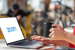 Person holding black mug and typing on laptop with Zoom logo on screen in a bright cafe setting