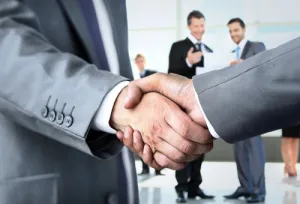 Close-up of business professionals shaking hands in a modern office with colleagues in the background.