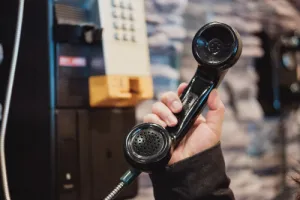 Hand holding a vintage black telephone handset near a wall-mounted payphone with a blurred background.