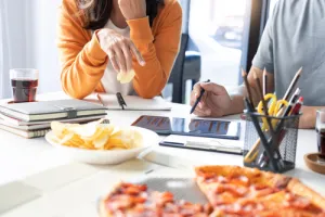 Two people working with tablets and notebooks at a table with pizza, chips, and drinks nearby.