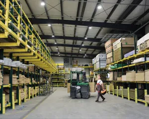 Worker walking in large warehouse aisle with stacked pallets, shelves, and a green forklift in center.