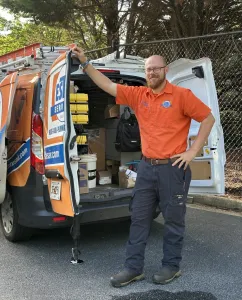 Estes Services Technician in orange uniform standing next to open service van with tools and equipment inside on sunny day.