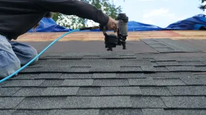 A person using a nail gun to install asphalt shingles on a roof under a clear blue sky.
