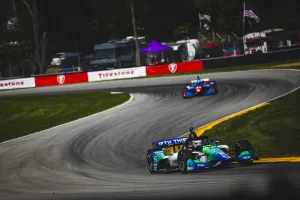 IndyCar race cars speed around a curved track with Firestone banners and spectators in the background.