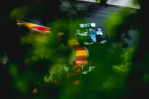 Two race cars speed through a curved section of a racetrack partially obscured by green foliage.