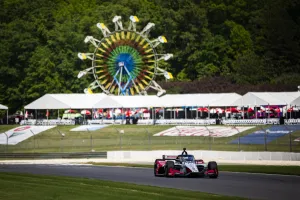 IndyCar race car speeding on track with colorful Ferris wheel and tents in background during event