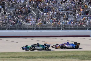 Two race cars speeding on a track with a large crowd watching from the grandstands in bright daylight.