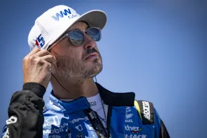 Race car driver in blue racing suit adjusts earpiece under clear blue sky, wearing white cap and sunglasses