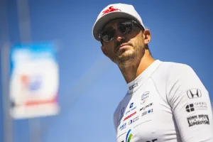 Race car driver wearing white racing suit and cap with sponsor logos against blue sky background