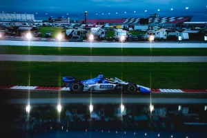 IndyCar race car speeding on track at dusk with RVs and grandstands in the background under floodlights