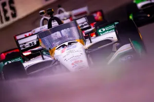 Close-up of a white IndyCar racing on track with visible sponsorship logos and helmet behind the windshield.