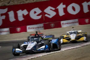 IndyCar race cars speeding on track with Firestone sponsor banners in the background at a racing event.