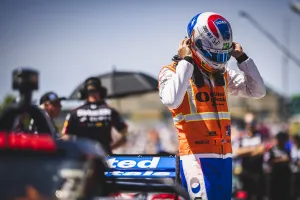 Race car driver adjusting helmet near blue race car in a motorsport event pit area.