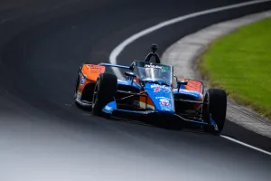 Blue and orange IndyCar racing at high speed on curved race track with green grass on side.