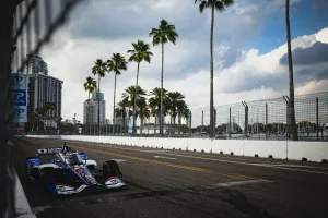IndyCar race car on a city street circuit lined with palm trees under a partly cloudy sky.