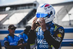 Race car driver adjusts helmet before a racing event with team members in blue uniforms in the background.
