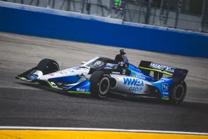 Blue and white IndyCar racing on an asphalt track with advertising logos and a blue wall background