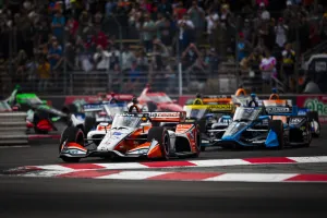 Open-wheel race cars compete closely on a curving racetrack with spectators in the background during a motorsport event.