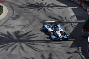 IndyCar racing car navigating a sharp turn on track with large palm tree shadows overhead in bright daylight