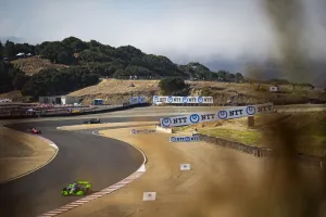 Race cars speeding on a winding track with dry hills and trees in the background at a motorsport event.