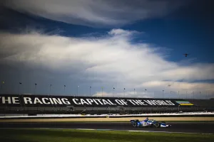 Blue and white racecar on track at raceway with grandstand reading The Racing Capital of the World under cloudy sky.