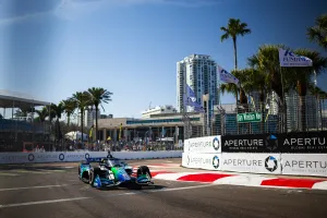 Race car speeding on urban track with palm trees, city buildings, and sponsors' banners in sunny weather.