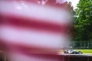 Blurred American flag in foreground with a blue and white race car speeding on a racetrack surrounded by green trees.