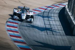 IndyCar race car navigating a sharp turn on a racetrack with red, white, and blue curbs under bright sunlight.
