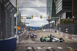 Open-wheel race cars competing on a city street circuit with barriers, fences, and traffic lights overhead.