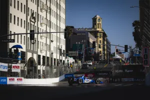 IndyCar race car speeds through city street circuit in sunny urban setting with historic clock tower.