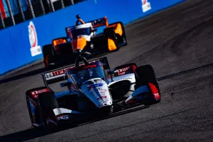 Two IndyCar race cars speeding on asphalt track with blue barrier and blurred background in bright sunlight.