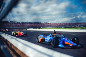 Blue and red Indy race cars speeding on a racetrack with a large crowd in the background under a cloudy sky.