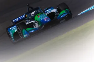 Green and blue IndyCar racing on track with sponsor logos and aerodynamic design under bright daylight.