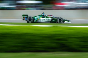 Green and white IndyCar racing at high speed on a racetrack with blurred background and green grass.