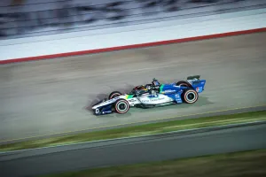 Blue and white IndyCar racing at high speed on a curved race track under stadium lights.