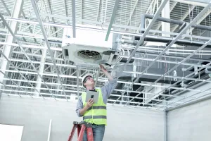 Technician inspecting ceiling HVAC unit, holding tablet, wearing safety vest and helmet in industrial building.