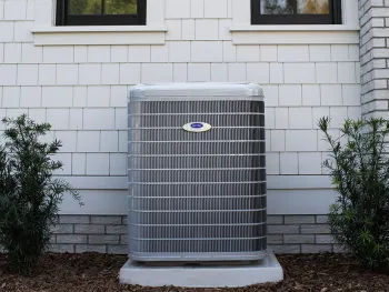A modern air conditioning unit installed outside a house with windows and greenery nearby.