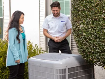 Technician in uniform explaining an air conditioning unit to a woman outside a home with greenery around.