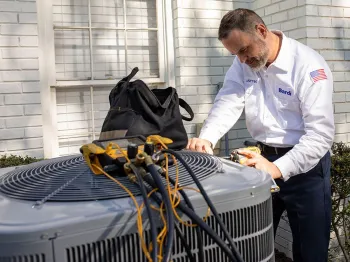 Technician servicing an air conditioning unit outside a white brick home with tools in hand.