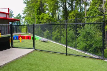 Black chain-link fence encloses a play area with artificial grass, colorful toys, and surrounding trees.