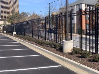 Black metal fencing alongside a parking lot, with pavement and greenery under a clear blue sky.