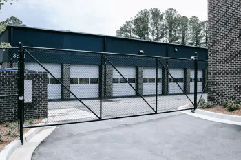 Black chain-link fence gate in front of a modern brick building with multiple garage doors and trees in the background.