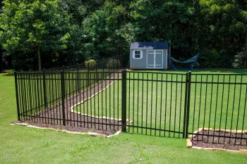 Black metal fence enclosing a grassy yard with a small gray shed and blue hammock under trees.