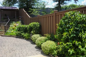 Neatly trimmed shrubs line a paved driveway beside a tall wooden fence under a sunny blue sky.
