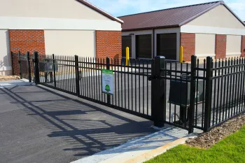 Black metal fence and gate securing a parking area outside brick and beige commercial buildings on a sunny day.