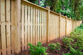 Long wooden fence with vertical planks surrounding a garden with green shrubs and trees in the background