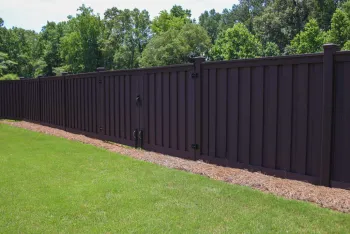 Long dark brown wooden fence with gate enclosing a grassy yard next to green trees in sunlight