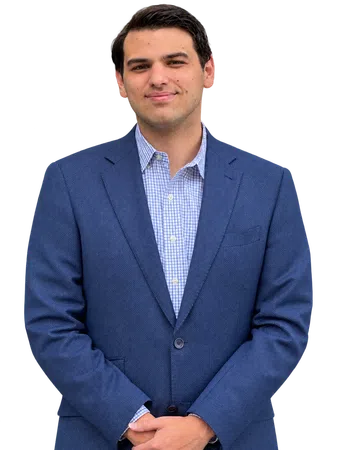 Confident young man in blue blazer and checkered shirt posing with hands clasped, smiling slightly.