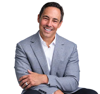 Smiling man wearing a gray checked blazer and white shirt sitting with arms crossed in a relaxed pose.
