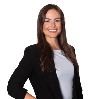 Smiling woman with long brown hair wearing a black blazer and light gray shirt posing confidently with hand in pocket